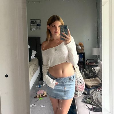 Alana Clements taking a mirror selfie wearing a white top with a blue skirt.